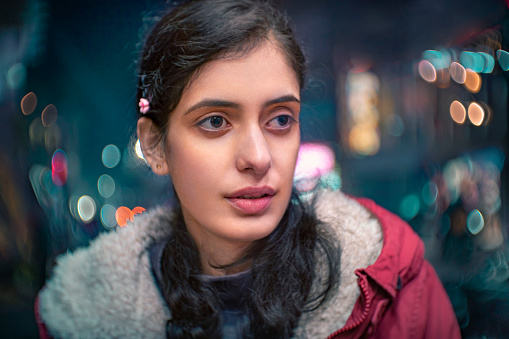 In this close-up, outdoor image, an Asian/Indian beautiful young woman stands on the side of a city street and looks away at night in winter. She is in a warm, red jacket.