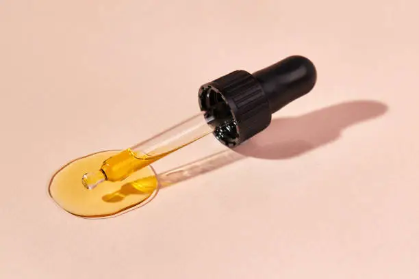 Close up of pipette with pouring liquid essential oil on beige background. Dropper with a falling drop of aromatherapy oil close-up