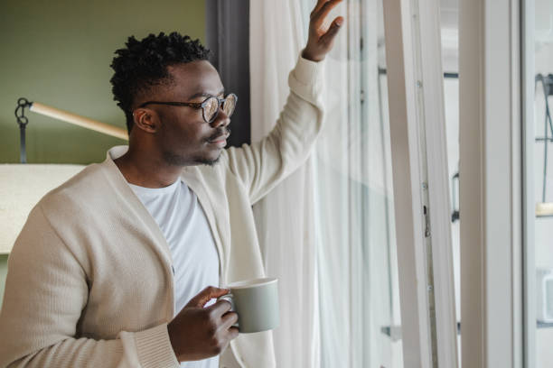 jeune homme afro-américain tenant une tasse de café et regardant par la fenêtre - homme mug regarder dehors photos et images de collection