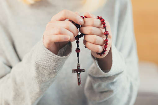 Close up photo of folded hands of a young woman holding a rosary, praying