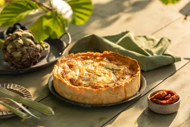 Golden organic healthy crispy pie quiche with artichokes, dried tomatoes and olives on the table, evening sunlight in the summer garden, soft selective focus