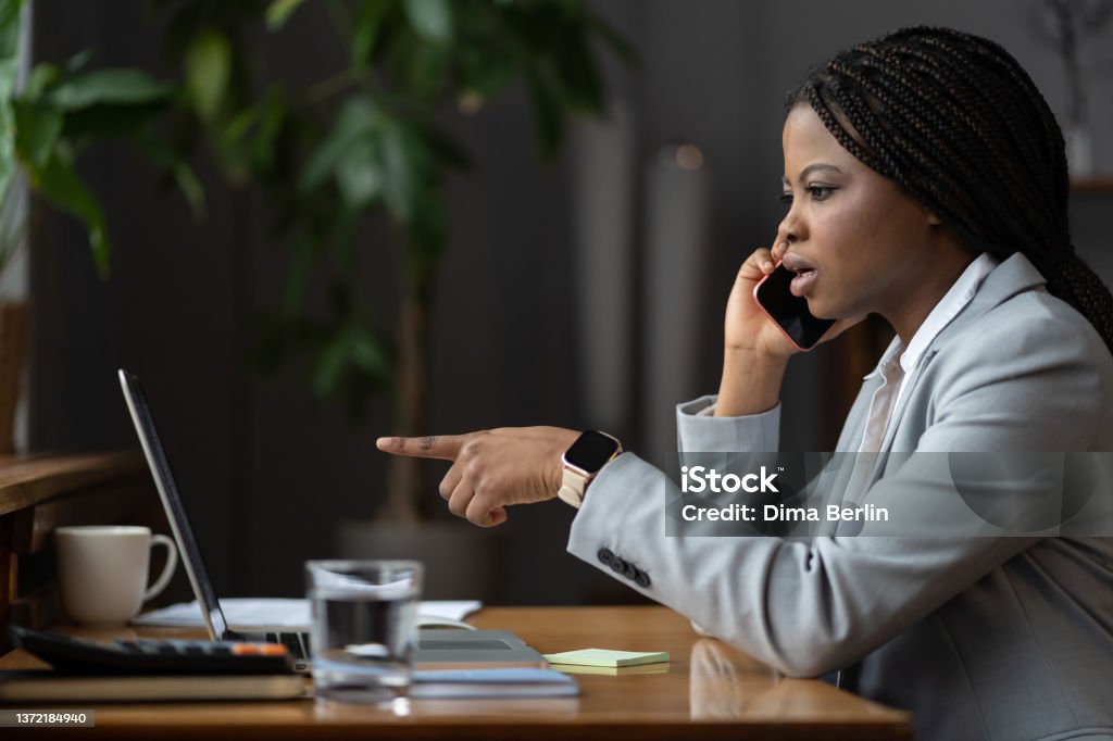 Shocked unhappy afro businesswoman talk on phone call point finger at laptop screen solving problem Frustrated african american businesswoman talk on phone with colleague ask for help point finger at laptop screen. Upset young woman hold smartphone consulting with coworker solving business problem Businesswoman Stock Photo