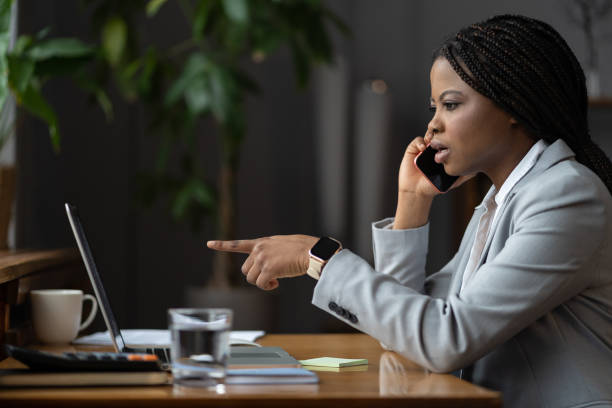 Shocked unhappy afro businesswoman talk on phone call point finger at laptop screen solving problem Frustrated african american businesswoman talk on phone with colleague ask for help point finger at laptop screen. Upset young woman hold smartphone consulting with coworker solving business problem melancholy computer support specialist stock pictures, royalty-free photos & images