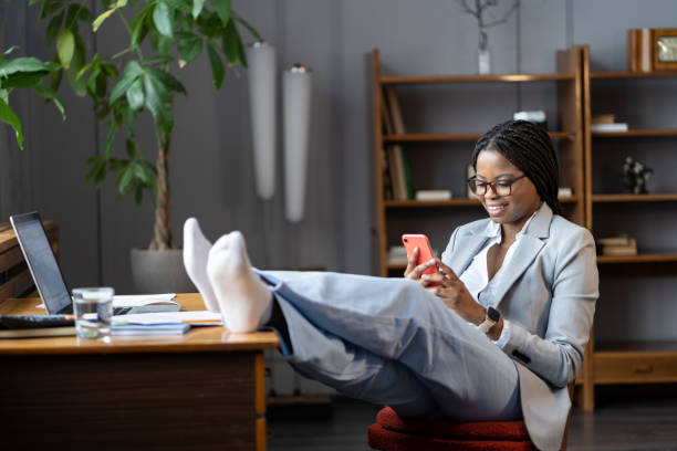 mujer de negocios afro relajada procrastina en el lugar de trabajo sentarse con los pies en el escritorio usar mensajes de teléfonos inteligentes - perder el tiempo fotografías e imágenes de stock