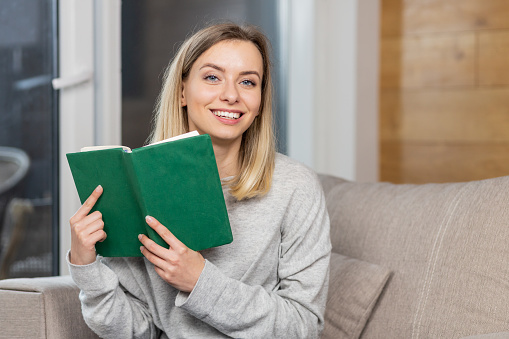 Portrait of a young beautiful woman reading a book, studying, sitting on the couch, resting, looking at the camera, smiling