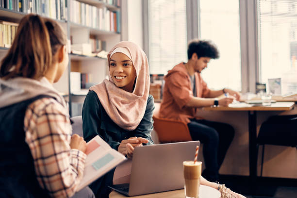 mulher muçulmana feliz falando com uma amiga enquanto estudavam juntas em uma biblioteca. - arabic language - fotografias e filmes do acervo