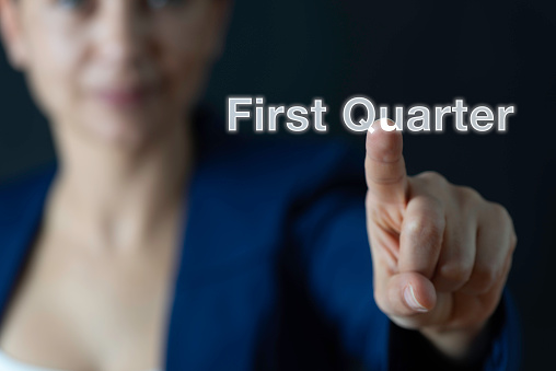 Businesswoman in dark room wearing navy blue suit is pointing at white First Quarter text in front of her.