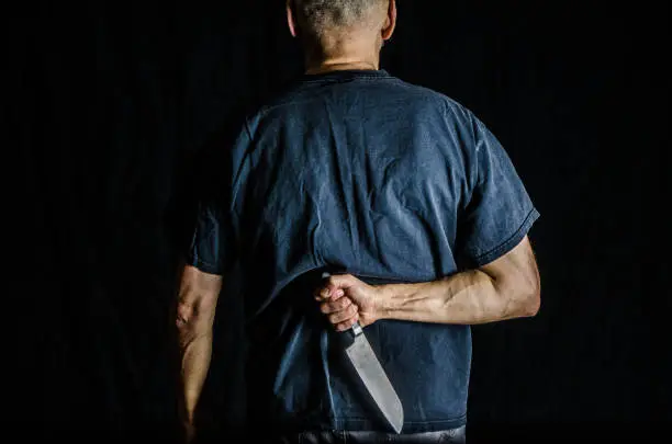 Man holding kitchen knife in his back, wearing black t-shirt, on black background