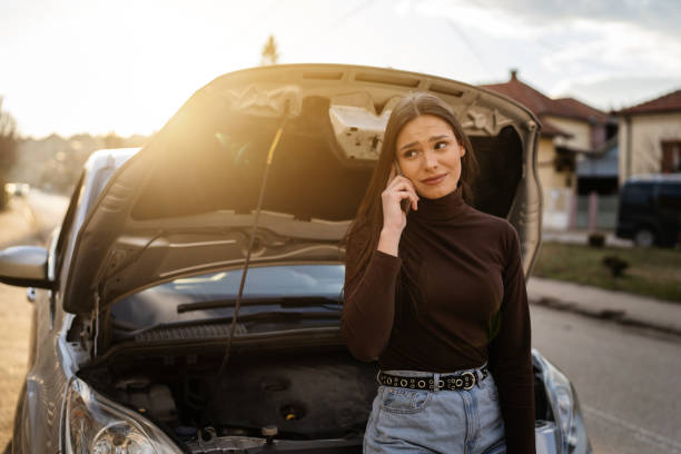 one young adult caucasian woman standing by her vehicle with open hood broken failed engine holding a phone calling towing service for help on the road roadside assistance concept autumn or spring day - vehicle breakdown stockfoto's en -beelden