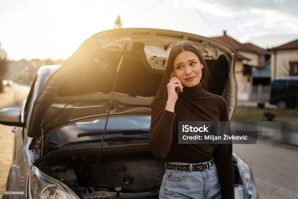 One young adult caucasian woman standing by her vehicle with open hood broken failed engine holding a phone calling towing service for help on the road Roadside assistance concept autumn or spring day Vehicle Breakdown Stock Photo