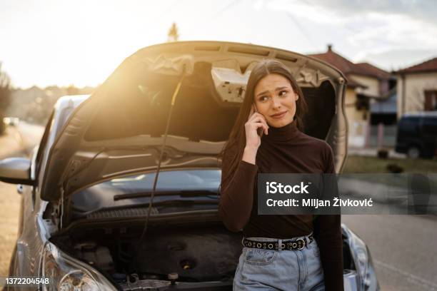 One Young Adult Caucasian Woman Standing By Her Vehicle With Open Hood Broken Failed Engine Holding A Phone Calling Towing Service For Help On The Road Roadside Assistance Concept Autumn Or Spring Day Stock Photo - Download Image Now