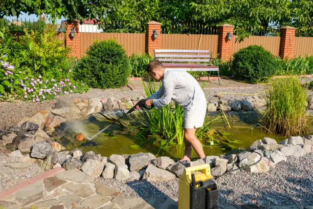 Young unrecognized caucasian guy cleans artificial garden pond bottom with high-pressure washer nozzle from mud and sludge. Spring and summer pond care work, back view