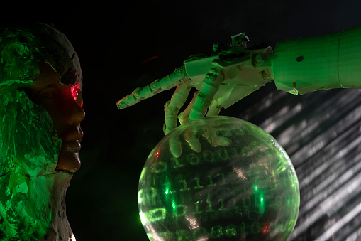 Humanoid robot hand pointing toward bust covered with clay in form of android. A red laser light is illuminating android eye. A sphere with binary code is seen under hand. Shot with a full frame mirrorless camera.