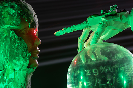 Humanoid robot hand pointing toward bust covered with clay in form of android. A red laser light is illuminating android eye. A sphere with binary code is seen under hand. Shot with a full frame mirrorless camera.