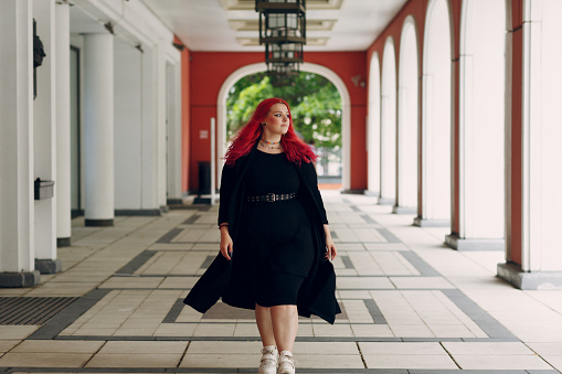 European plus size woman walking outdoor street city. Young red pink haired body positive girl.