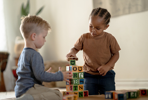 Two Sweet little preschool aged children play together on the floor with a stack of wooden building blocks.  The boys are both dressed casually and working together as they focus on stacking the colorful letter blocks into a tower.