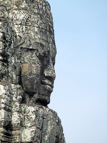 Close-up of the profile of a large face carved in stone at the famous Bayon temple, Angkor Wat, Siem Reap, in Cambodia. Face is set to the left of frame, blue sky background to the right.
