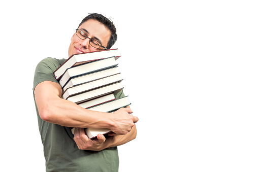 Nerdy man in glasses smiling and hugging pile of favorite books with closed eyes while standing against white background