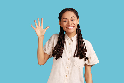 Cheerful friendly woman with black dreadlocks waving palm in hello gesture, meets someone at street, smiles positively, wearing white shirt. Indoor studio shot isolated on blue background.