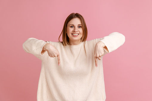 Portrait of attractive happy blond woman pointing down place for commercial idea, looking at camera with toothy smile, wearing white sweater. Indoor studio shot isolated on pink background.