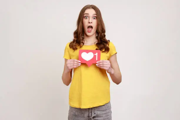 Photo of Like and follow my blog! Shocked blogger young woman in yellow T-shirt holding social media Heart button, astonishing of amount of followers.