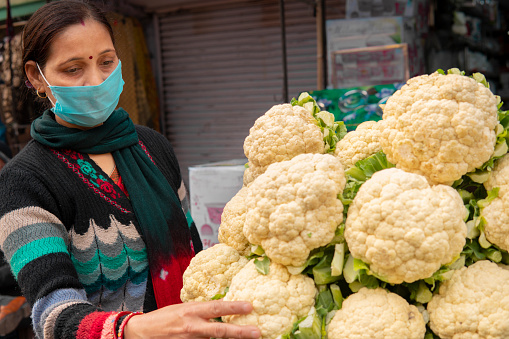 Panoramic image of Indian mature woman wearing protective face mask and buying fresh green vegetables from outdoor market during the third wave of coronavirus, COVID-19 pandemic outbreak in Indian.