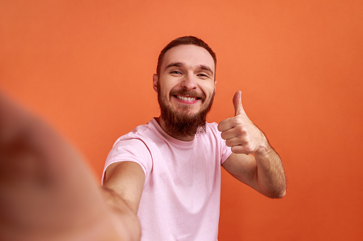 Portrait of positive handsome bearded man making selfie or streaming, showing thumb up POV, point of view of photo, wearing pink T-shirt. Indoor studio shot isolated on orange background.