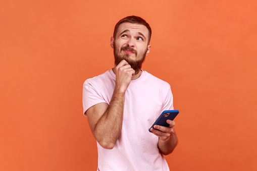 Portrait of man holding chin, standing with smartphone in hand, contemplating about software updating, choosing suitable tariffs, wearing pink T-shirt. Indoor studio shot isolated on orange background
