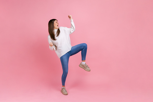 Full length portrait of extremely happy female celebrating her success, standing with clenched fists, wearing white casual style sweater. Indoor studio shot isolated on pink background.