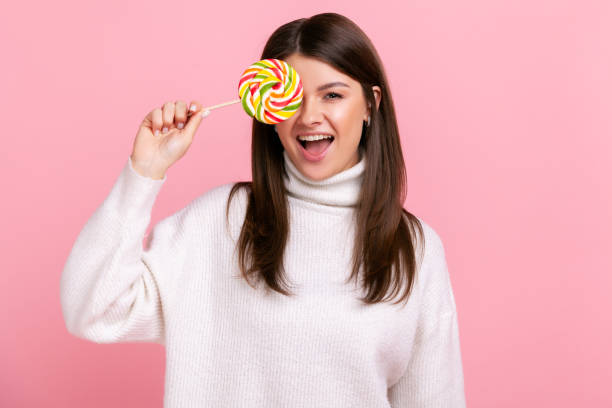 Portrait of brunette female covering eye with big tasty candy, looking at camera, showing tongue out Portrait of brunette female covering eye with big tasty candy, looking at camera, showing tongue out, wearing white casual style sweater. Indoor studio shot isolated on pink background. eye candy stock pictures, royalty-free photos & images