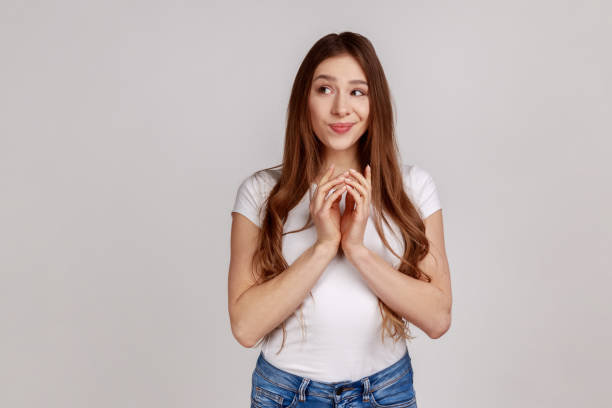 Scheming devious woman clasping hands, thinking over tricky plan, having sly cunning idea to prank. Scheming devious attractive woman clasping hands and thinking over tricky plan, having sly cunning idea to prank, wearing white T-shirt. Indoor studio shot isolated on gray background. sideways glance stock pictures, royalty-free photos & images