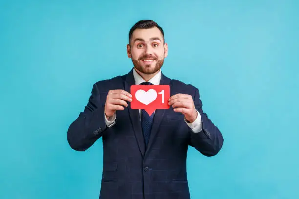 Photo of Satisfied man with beard wearing official style suit holding heart icon, recommending to follow and share blog with interesting content.