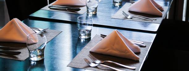 serviettes blanches pliées sur tapis de table, fourchettes de couteaux en acier inoxydable et verres à boire en cristal sur table en bois noir - spoon napkin silverware white photos et images de collection