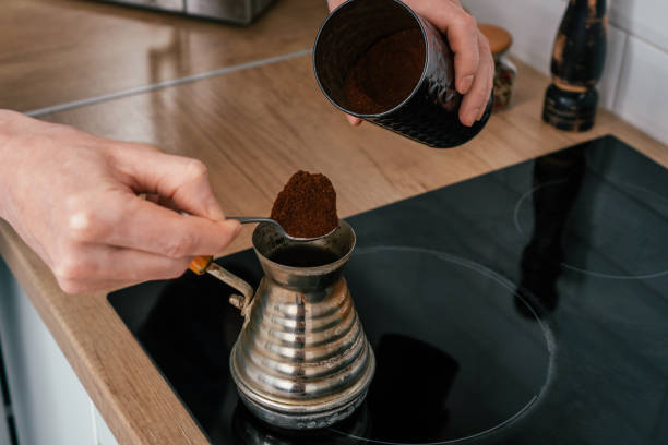 Close up of hands of unrecognizable man putting full heaped tea spoon of ground coffee from round tin in copper cezve. Close up of hands of unrecognizable man, putting full heaped tea spoon of natural ground coffee from round glossy tin in copper cezve on black electric cooker for brewing near spice grinder in kitchen cezve stock pictures, royalty-free photos & images