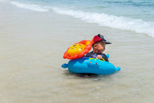 Happy little Asian child on the tropical beach. Boy playing swim ring with sea wave on the ocean in the summer holiday. Kid with inflatable ring in nature family vacation. Children sit at the seashore.