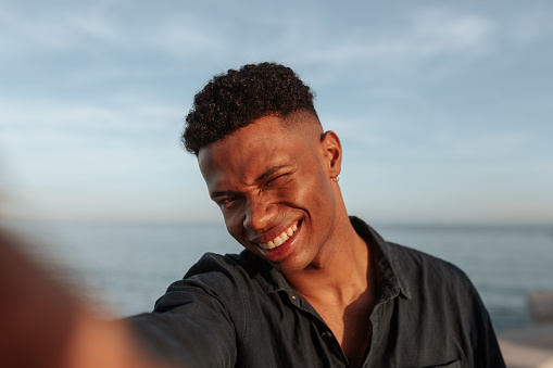 Young black man winking at the camera while taking a selfie. Cheerful young man smiling and having a good time by the seaside. Outdoorsy young man capturing his happy moments.