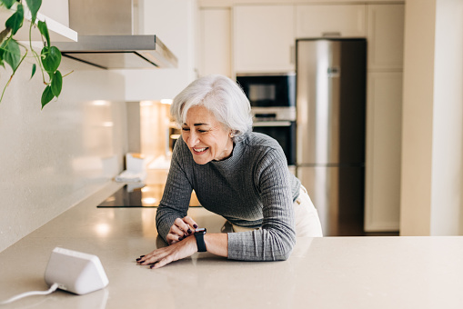 Senior woman smiling happily while using smart devices in her kitchen. Cheerful elderly woman using a home assistant to perform tasks at home.