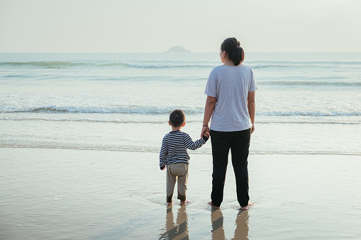 Rear view of happy Asian mother and her little son bonding together on the tropical beach. Family of Mom walking with child while on vacation beautiful sea in summer day. Copy space. Holiday concept