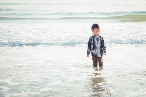 Happy little Asian child smiling on the tropical beach. Boy playing sea waves while on vacation beautiful ocean in the summer holiday. Kid in nature at sunset. Children walking at seashore. Copy space