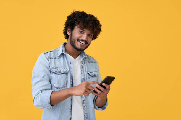 istock Smiling indian young man using cell phone isolated on yellow background. 1372303815