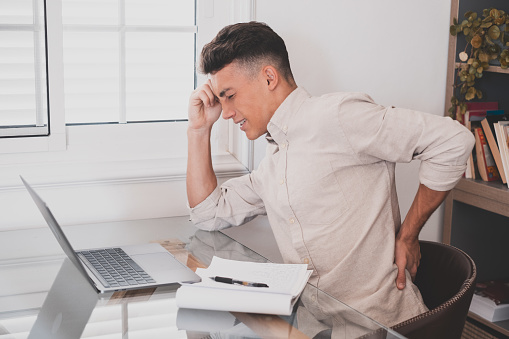 Close up rear view stressed young man touching lower back feeling discomfort, suffering from sudden pain due to sedentary lifestyle or long computer overwork in incorrect posture at home office.