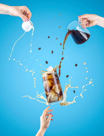 Woman hand support fly glass of tasty brew cold coffee and ice with splash, women hands is pouring homemade sour cream from small jar and coffee from kettle to glassful, ingredients falling
