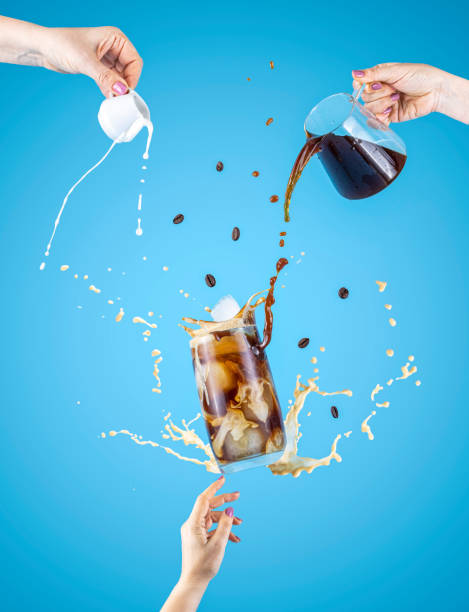 Woman hand support fly glass of tasty brew cold coffee and ice with splash, women hands is pouring homemade sour cream from small jar and coffee from kettle to glassful stock photo