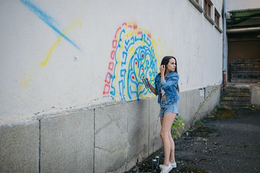 Beautiful young woman paints graffiti on concrete wall with aerosol paint can