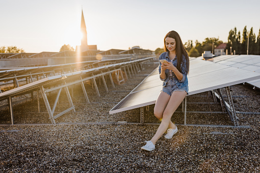Beautiful young woman text messaging on smartphone while sitting on solar panels at power station