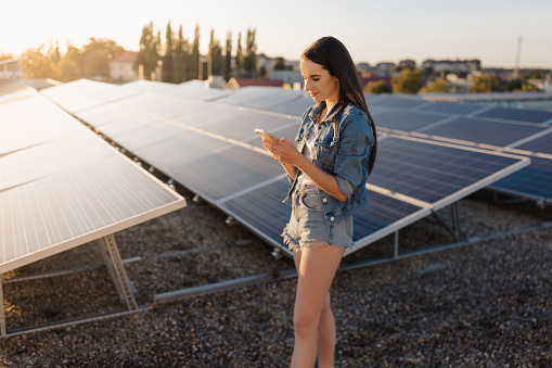 Beautiful young woman text messaging on smartphone while standing beside solar panels at power station