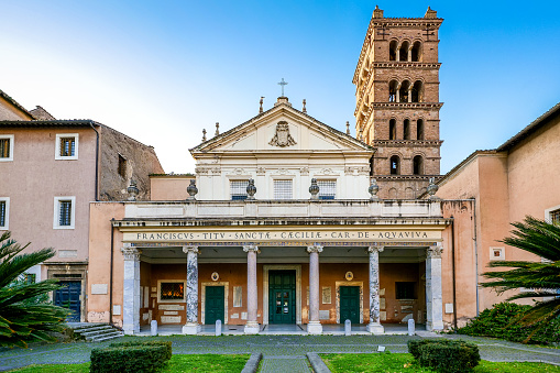 The facade and the medieval bell tower of the Basilica di Santa Cecilia (St. Cecilia Basilica), in the ancient Trastevere district, in the historic heart of Rome. According to legend, this church was built starting from the 5th century in the place where St. Cecilia was martyred, in 220 AD. The church took on its current Baroque style after some transformations made in the 16th century. Inside, under the main altar, you can admire the marble statue of St. Cecilia made in 1599 by the sculptor Stefano Maderno, which depicts the body of the saint covered by a veil, as it was in her original tomb present in the ancient perimeter of the church. Trastevere is an iconic district of the Eternal City, due to the presence of countless artistic and historical treasures, monuments and ancient Romanesque and Baroque churches, but also for its squares and alleys to be explored freely, where it is easy to find typical restaurants, pubs, small shops of artisans and scenes of daily life with the original Roman soul. In 1980 the historic center of Rome was declared a World Heritage Site by Unesco. Super wide angle image in high definition format.