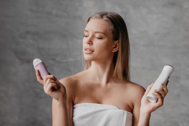 Young woman choosing between roller and spray deodorants , wearing towel while standing in bathroom after shower stock photo