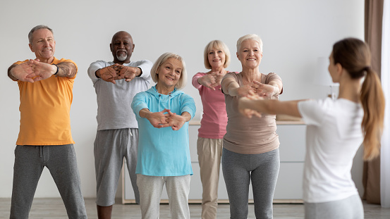Sporty multiracial group of elderly people having fitness class at nursing home, training with female instructor, standing in a row, stretching hands and smiling, enjoying physical activity, panorama