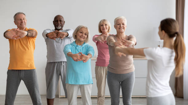 Sporty elderly people having fitness class, training with instructor Sporty multiracial group of elderly people having fitness class at nursing home, training with female instructor, standing in a row, stretching hands and smiling, enjoying physical activity, panorama gym group class stock pictures, royalty-free photos & images